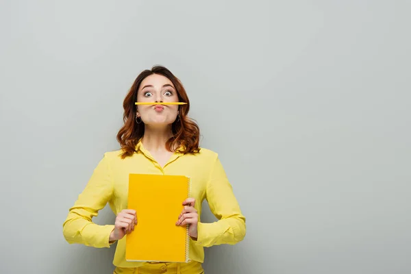 Cheerful woman with pencil between lips and nose looking at camera on grey — Stock Photo