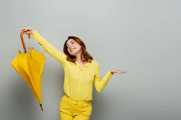 Fröhliche Frau in gelbem Hemd mit offenem Arm und gelbem Regenschirm auf grau — Stockfoto