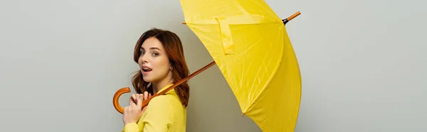 Flirty woman sticking out tongue while posing with yellow umbrella on grey, banner — Stock Photo