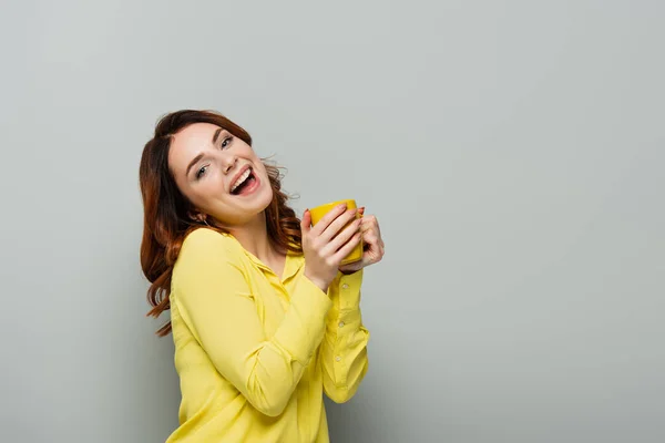Excité femme regardant caméra tout en tenant tasse jaune sur gris — Photo de stock