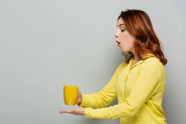 Donna scioccata in camicetta gialla contenente tazza di bevanda calda su grigio — Foto stock