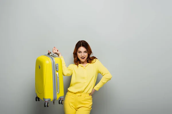 Femme excitée tenant une valise jaune debout avec la main sur la hanche sur gris — Photo de stock