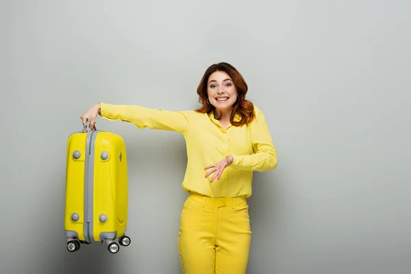 Femme joyeuse souriant à la caméra tout en pointant vers la valise jaune sur gris — Photo de stock