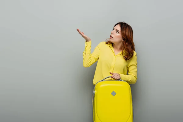 Шокована жінка вказує рукою, тримаючи жовту валізу на сірому — стокове фото