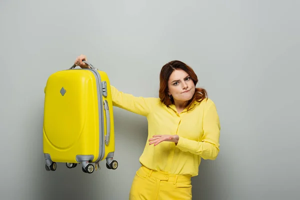Donna scoraggiata che indica la valigia gialla mentre guarda la fotocamera sul grigio — Foto stock