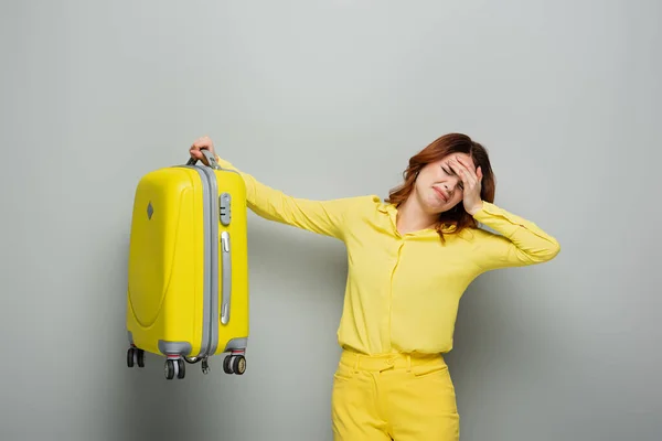 Femme insatisfaite debout avec les yeux fermés et touchant le front tout en tenant la valise sur gris — Photo de stock