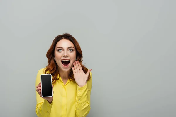 Verblüffte Frau blickt in Kamera, während sie Handy mit leerem Bildschirm in grau isoliert hält — Stockfoto