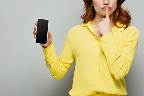 Ausgeschnittene Ansicht einer Frau, die Schweigegegeste zeigt, während sie ihr Handy mit leerem Bildschirm auf grau hält — Stockfoto