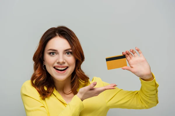 Cheerful woman looking at camera while pointing at credit card isolated on grey — Stock Photo