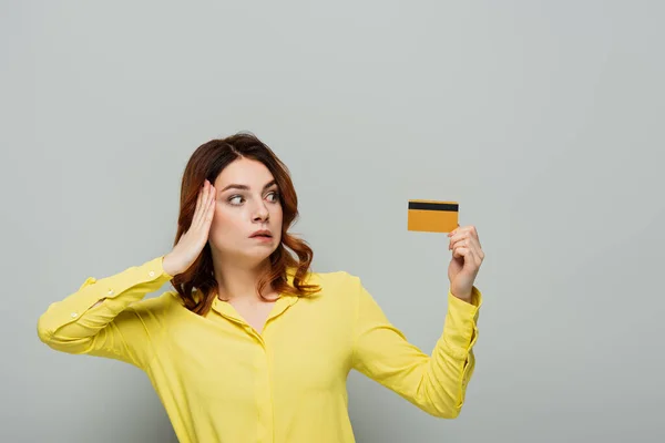 Femme perplexe touchant la tête tout en tenant la carte de crédit sur gris — Photo de stock