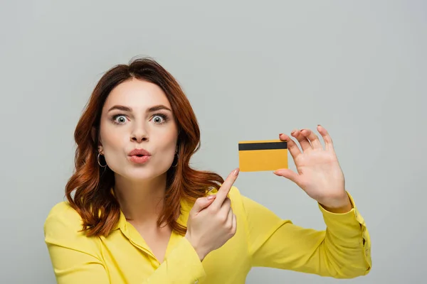 Surprised woman pouting lips while pointing with finger at credit card isolated on grey — Stock Photo