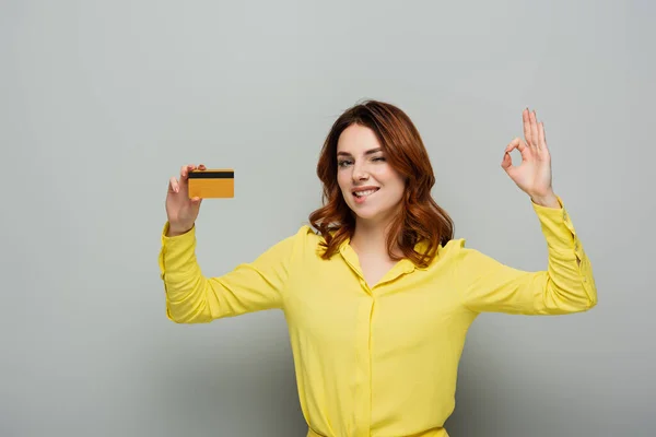 Cheerful woman holding credit card while showing ok gesture on grey — Stock Photo