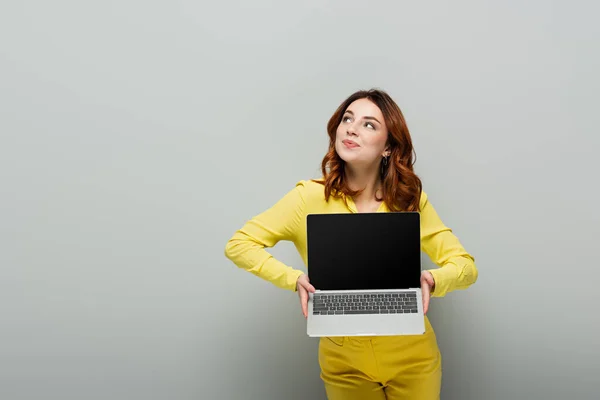 Sorrindo mulher olhando embora enquanto segurando laptop com tela em branco no cinza — Fotografia de Stock