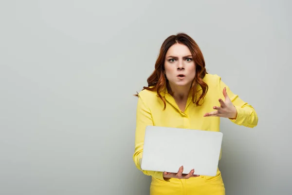 Роздратована жінка жестикулює, тримаючи ноутбук на сірому — стокове фото
