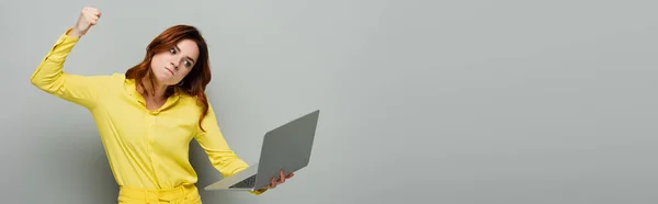 Femme irritée tenant ordinateur portable tout en se tenant debout avec le poing serré sur gris, bannière — Photo de stock