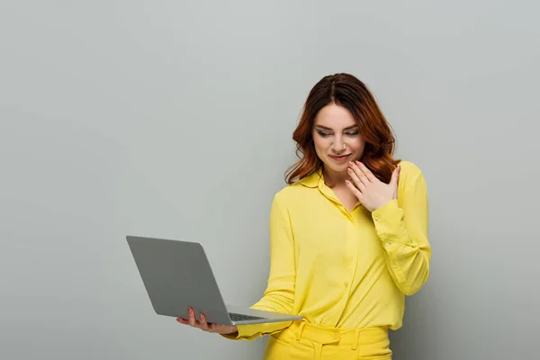 Mujer sonriente sosteniendo la mano cerca de la cara mientras sostiene el ordenador portátil en gris - foto de stock