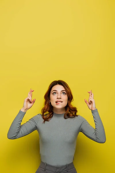 Mulher esperançosa segurando dedos cruzados enquanto olha para cima no amarelo — Fotografia de Stock