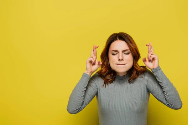 Mujer preocupada con los ojos cerrados sosteniendo los dedos cruzados mientras está de pie en amarillo - foto de stock