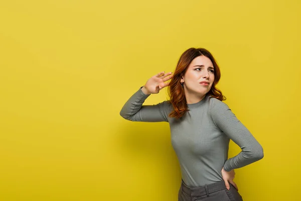 Displeased woman with curly hair standing with hand on hip while listening on yellow — Stock Photo