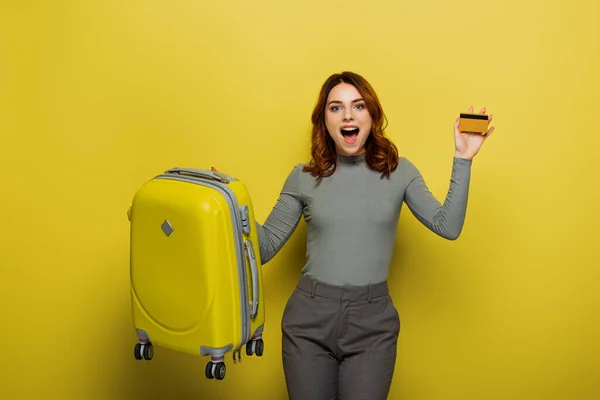 Verblüffte Frau mit lockigem Haar, die Gepäck und Kreditkarte auf gelb hält — Stockfoto