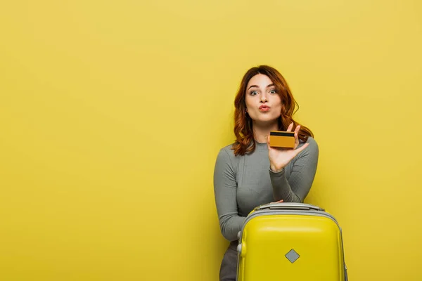 Mujer con el pelo rizado haciendo pucheros labios mientras sostiene el equipaje y la tarjeta de crédito en amarillo - foto de stock