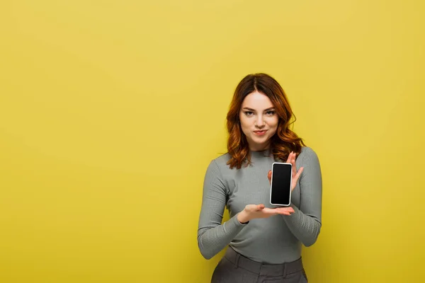 Fröhliche Frau mit lockigem Haar hält Smartphone mit leerem Bildschirm auf gelb — Stockfoto