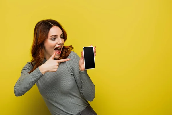 Mulher espantada com cabelo encaracolado apontando com o dedo para smartphone com tela em branco no amarelo — Fotografia de Stock