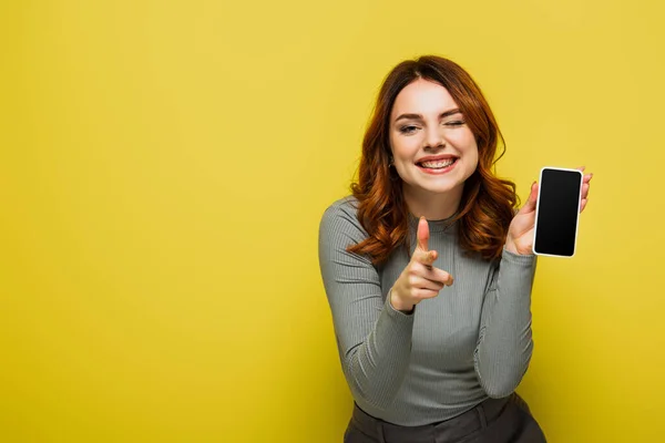 Mujer alegre sosteniendo teléfono inteligente con pantalla en blanco y apuntando con el dedo mientras mira a la cámara en amarillo - foto de stock