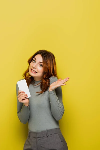 Ahnungslose junge Frau mit lockigem Haar hält Smartphone in der Hand und gestikuliert auf Gelb — Stockfoto