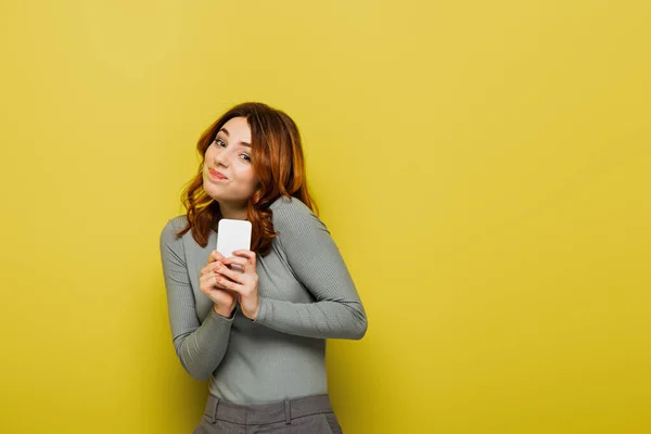 Verwirrte junge Frau mit lockigem Haar hält Smartphone in der Hand und blickt in Kamera auf gelb — Stockfoto