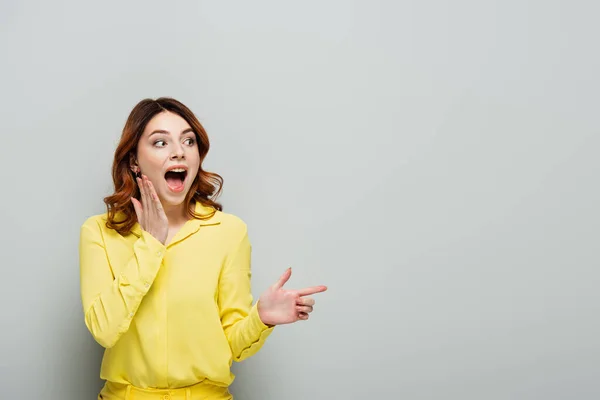 Jeune femme étonnante en chemise jaune pointant du doigt et regardant loin sur gris — Photo de stock