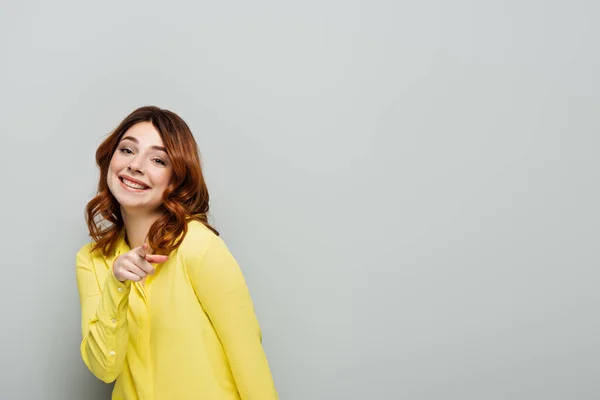 Молода позитивна жінка в жовтій сорочці вказує пальцем, дивлячись на камеру на сірій — стокове фото