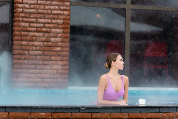 Femme en maillot de bain regardant loin dans la piscine extérieure près de tasse de café sur la surface de pierre — Photo de stock