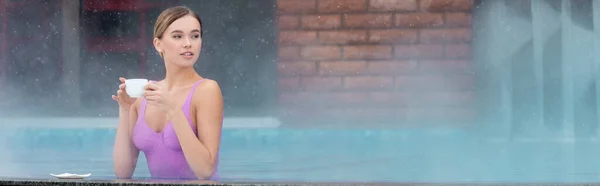 Jeune femme en maillot de bain tenant une tasse de café dans la piscine extérieure, bannière — Photo de stock