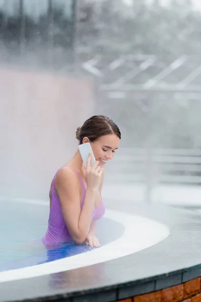 Femme gaie en maillot de bain parlant sur smartphone tout en se baignant dans la piscine extérieure — Photo de stock