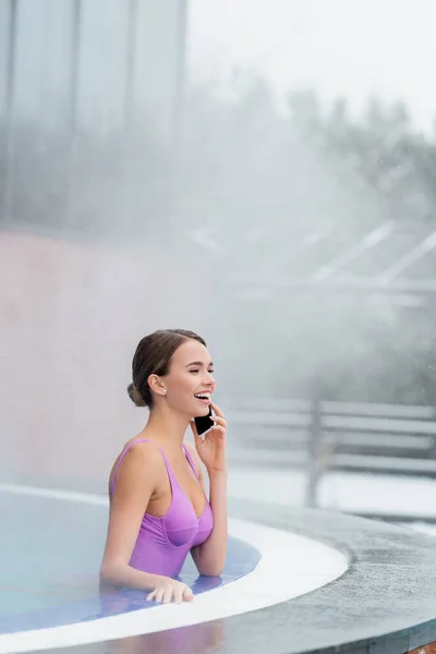 Femme heureuse en maillot de bain parlant sur smartphone tout en riant et en se baignant dans la piscine extérieure — Photo de stock
