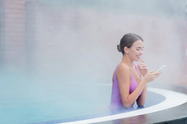 Femme heureuse en maillot de bain tenant smartphone tout en se baignant dans la piscine extérieure dans le centre de spa — Photo de stock