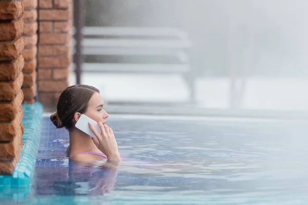 Vue latérale de la femme en maillot de bain parlant sur smartphone tout en se baignant dans la piscine extérieure — Photo de stock