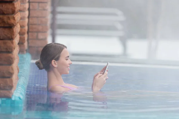 Seitenansicht einer glücklichen Frau, die ihr Smartphone beim Baden im Freibad benutzt — Stockfoto