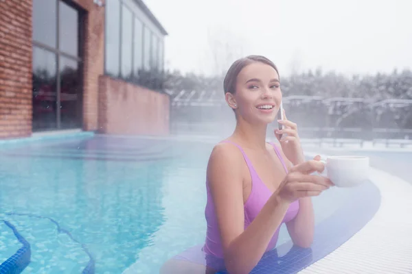 Allegra giovane donna che parla su smartphone e tiene in mano la tazza mentre fa il bagno nella piscina termale all'aperto — Foto stock