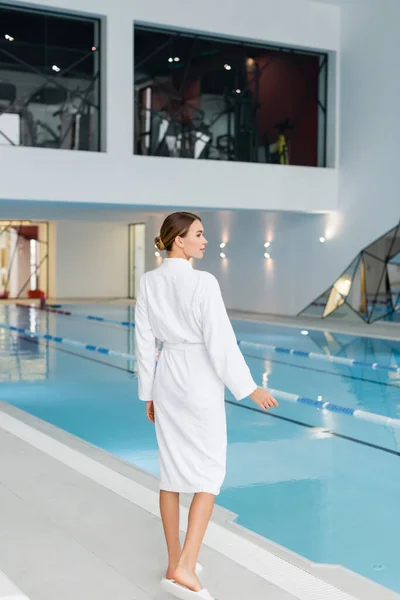 Longueur pleine de jeune femme en peignoir blanc debout près de la piscine dans le centre de spa — Photo de stock