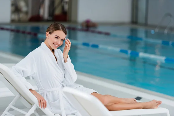 Веселая молодая женщина в белом халате смотрит в сторону, отдыхая на шезлонге возле бассейна — стоковое фото