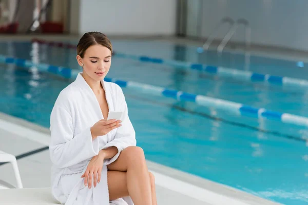 Young woman in white bathrobe sitting on deck chair and holding smartphone in spa center — Stock Photo