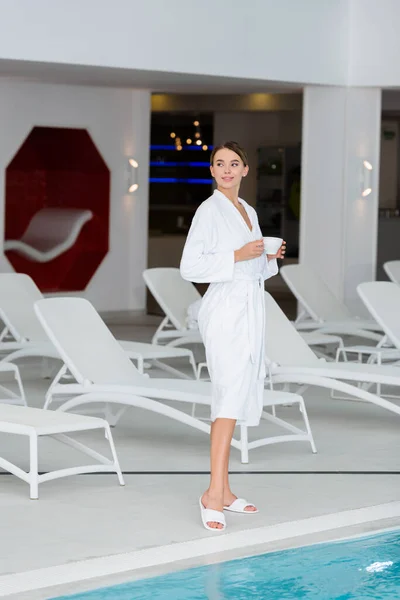 Femme positive en peignoir et pantoufles tenant tasse près de chaises longues et piscine dans le centre de spa — Photo de stock