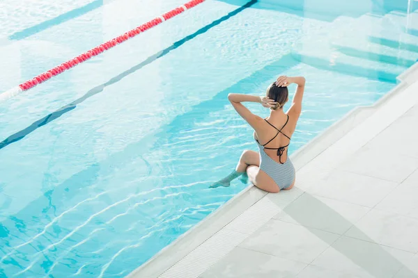 Задний вид молодой женщины в купальнике, регулирующей волосы, сидя возле бассейна в спа-центре — стоковое фото