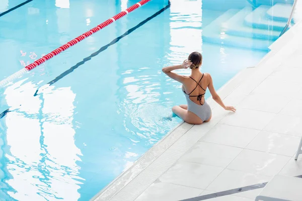 Vista trasera de la joven mujer ajustando el cabello mientras está sentada cerca de la piscina en el centro de spa - foto de stock