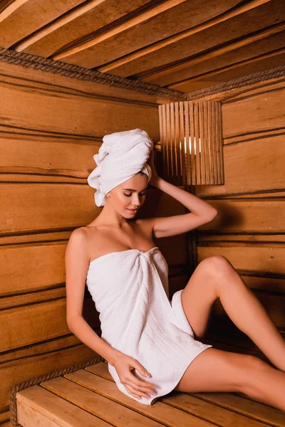 Young woman wrapped in towels sitting on seat in wooden sauna — Stock Photo