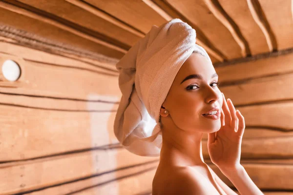 Lächelnde Frau mit Handtuch auf dem Kopf blickt in Sauna auf verschwommenem Hintergrund in die Kamera — Stockfoto