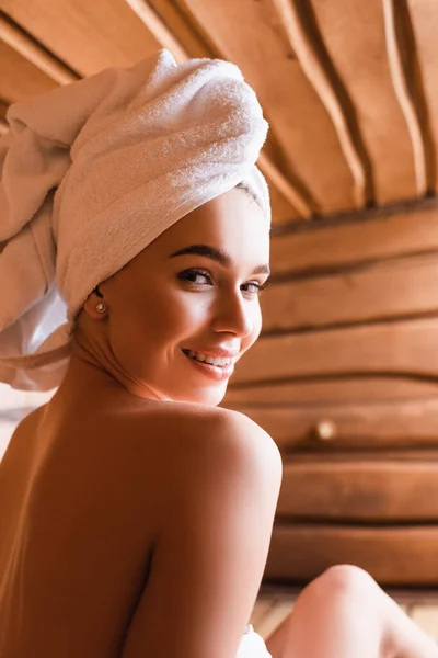Donna con asciugamano sulla testa sorridente alla macchina fotografica in sauna su sfondo sfocato — Foto stock