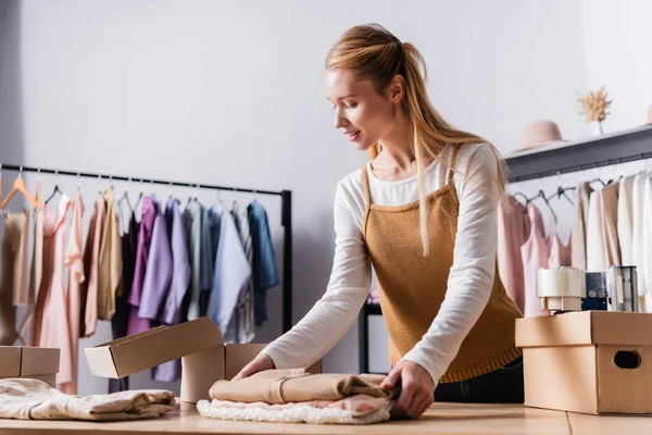 Mujer de negocios recogiendo orden cerca de cajas de cartón en sala de exposición cerca de perchas sobre fondo borroso - foto de stock